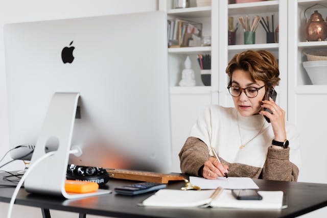 person-at-desk-with-monitor-talking-on-phone-and-writing-notes-on-notepad person-at-desk-with-monitor-talking-on-phone-and-writing-notes-on-notepad